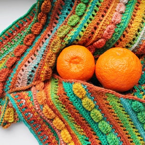May include: A colorful crocheted blanket with a striped pattern in shades of orange, green, and brown. Two whole oranges are resting on the blanket.