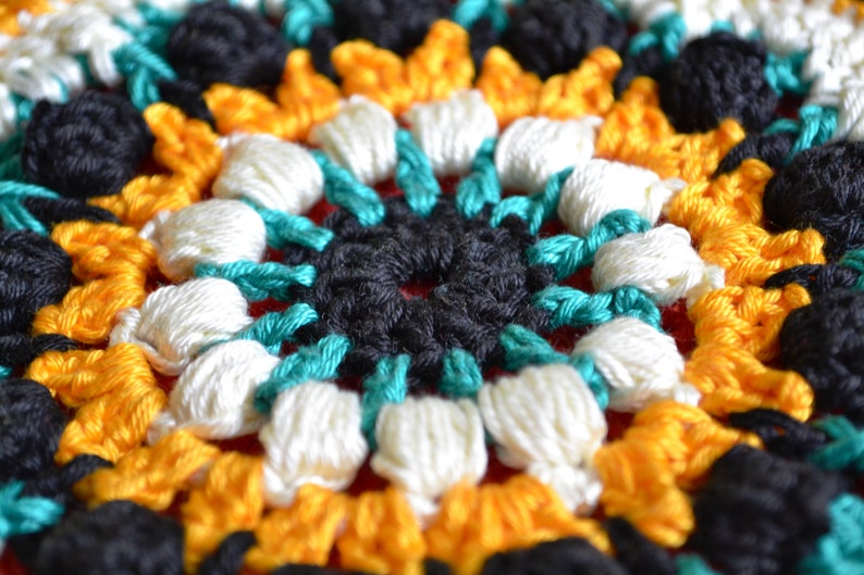 May include: Close-up of a crocheted mandala with a central black circle surrounded by turquoise, white, and yellow yarn.