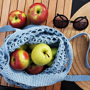 May include: A blue crocheted mesh bag filled with green and red apples. A straw hat and sunglasses are on a wooden surface next to the bag.