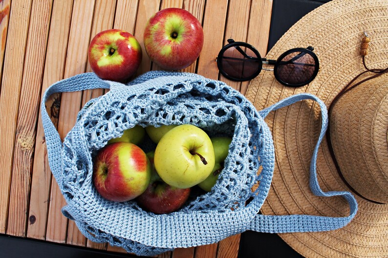May include: A blue crocheted mesh bag filled with green and red apples. A straw hat and sunglasses are on a wooden surface next to the bag.