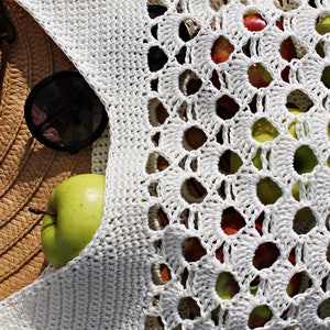 May include: A white crocheted bag with large holes, filled with green and red apples. A straw hat and sunglasses are partially visible in the background.