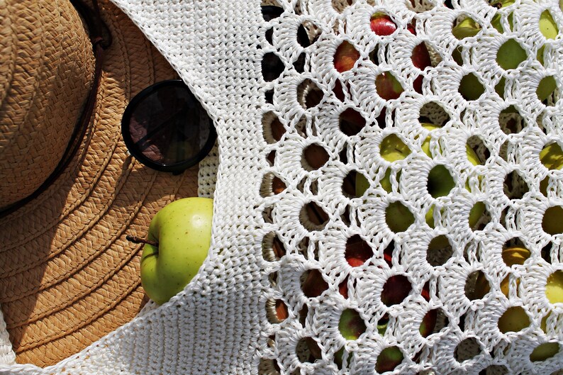May include: A white crocheted bag with large holes, filled with green and red apples. A straw hat and sunglasses are partially visible in the background.