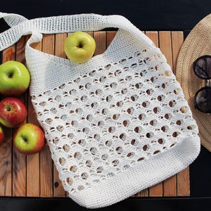 May include: A white crocheted tote bag with a long strap, filled with green and red apples. The bag is sitting on a wooden surface next to a straw hat and sunglasses.
