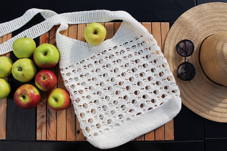 May include: A white crocheted tote bag with a long strap, filled with green and red apples. The bag is sitting on a wooden surface next to a straw hat and sunglasses.