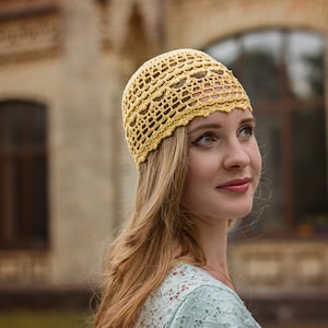 Woman in a handmade light yellow cotton summer hat against an urban landscape. The hat features an openwork design with floral elements.