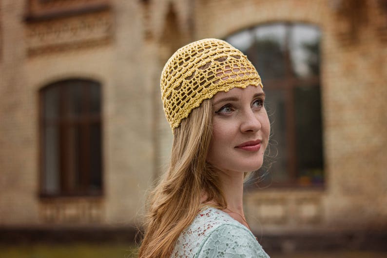 Woman in a handmade light yellow cotton summer hat against an urban landscape. The hat features an openwork design with floral elements.