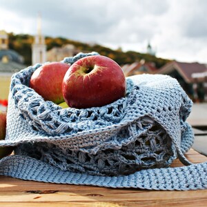 May include: A blue crocheted bag with a long strap, filled with red apples. The bag is sitting on a wooden table.