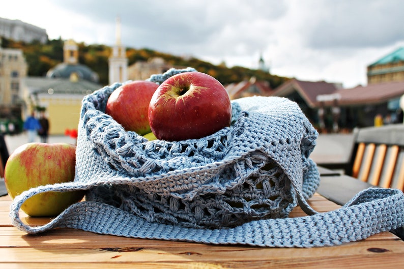 May include: A blue crocheted bag with a long strap, filled with red apples. The bag is sitting on a wooden table.