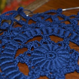 May include: A close-up of a blue crocheted doily with a delicate, intricate pattern. The doily is made with a single strand of yarn and has a circular shape.