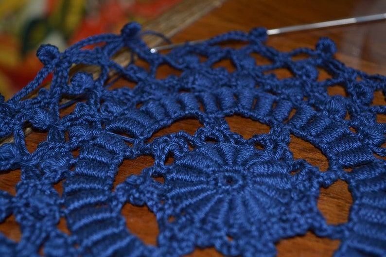 May include: A close-up of a blue crocheted doily with a delicate, intricate pattern. The doily is made with a single strand of yarn and has a circular shape.