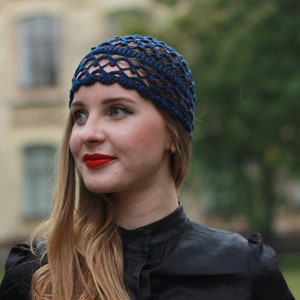 May include: A woman with long blonde hair is wearing a dark blue crochet hat with a scalloped edge. She is wearing a black mock neck shirt and has red lipstick on.