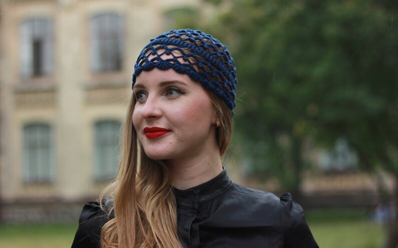 May include: A woman with long blonde hair is wearing a dark blue crochet hat with a scalloped edge. She is wearing a black mock neck shirt and has red lipstick on.