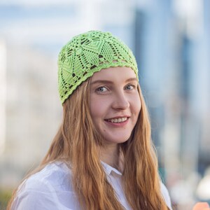 May include: A woman with long, red hair is wearing a light green crocheted hat. She is smiling and looking at the camera.