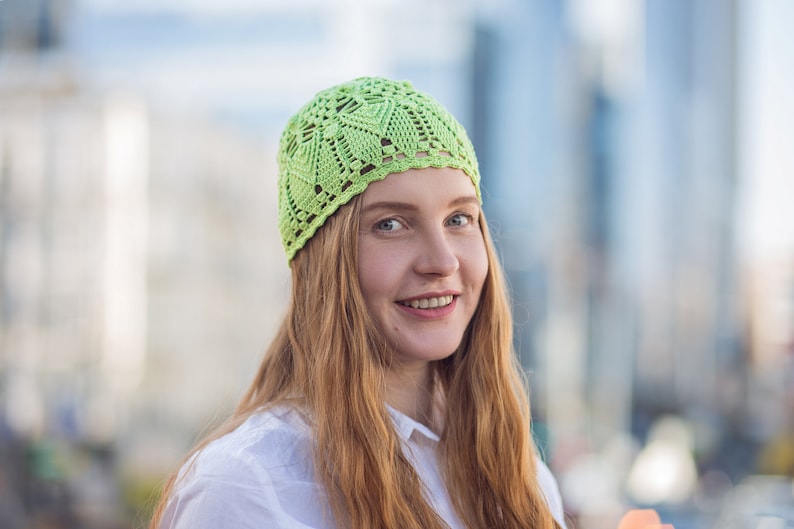May include: A woman with long, red hair is wearing a light green crocheted hat. She is smiling and looking at the camera.