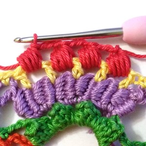 May include: Close-up of a crocheted rainbow mandala with a pink crochet hook. The hook is holding a red yarn and is in the process of creating a new row of crochet stitches.