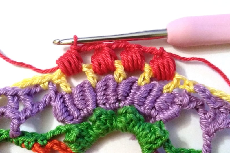 May include: Close-up of a crocheted rainbow mandala with a pink crochet hook. The hook is holding a red yarn and is in the process of creating a new row of crochet stitches.