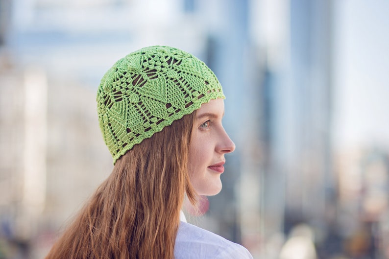 May include: A green crocheted hat with a delicate pattern. The hat is worn by a person with long, red hair.