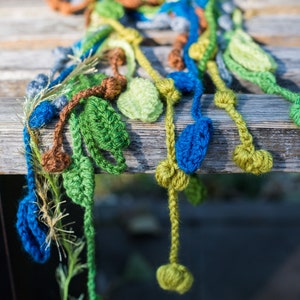 May include: A crocheted scarf with green, blue, and brown leaves hanging from it. The scarf is made of yarn and has a textured look.