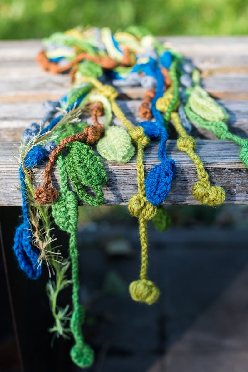 May include: A crocheted scarf with green, blue, and brown leaves hanging from it. The scarf is made of yarn and has a textured look.