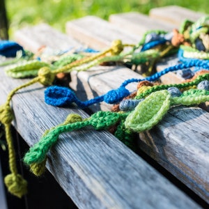 May include: A close-up of a wooden bench with a colorful crocheted garland made of green, blue, and brown yarn. The garland is made of leaves and beads.