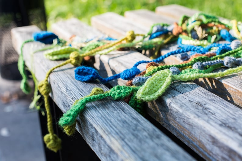 May include: A close-up of a wooden bench with a colorful crocheted garland made of green, blue, and brown yarn. The garland is made of leaves and beads.