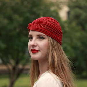May include: A woman with long, blonde hair is wearing a red crocheted hat. The hat has a unique design with a small bow on the side. She is looking off to the side with a slight smile.