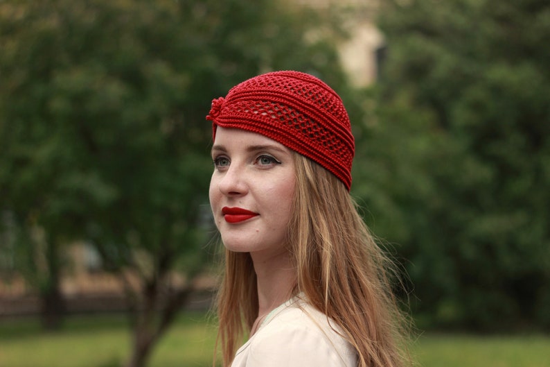 May include: A woman with long, blonde hair is wearing a red crocheted hat. The hat has a unique design with a small bow on the side. She is looking off to the side with a slight smile.