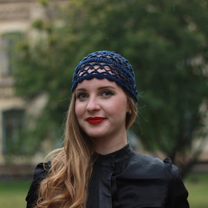 May include: A woman with long blonde hair wearing a blue crocheted hat and a black mock neck shirt.