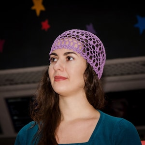 May include: A woman with long brown hair wearing a purple crochet hat and a teal blue shirt.