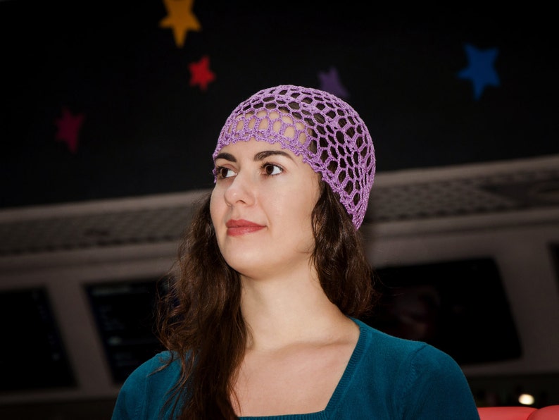 May include: A woman with long brown hair wearing a purple crochet hat and a teal blue shirt.