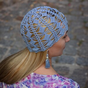 A woman stands partially turned, displaying an exquisite handmade crocheted hat with a lace pattern.
