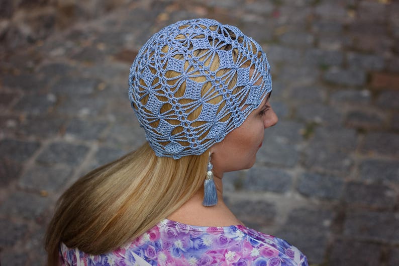 A woman stands partially turned, displaying an exquisite handmade crocheted hat with a lace pattern.