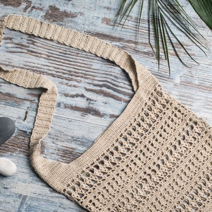May include: A beige crocheted tote bag with a long strap, lying on a light blue wooden surface with stones and a green leaf.
