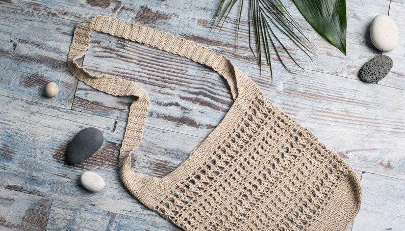 May include: A beige crocheted tote bag with a long strap, lying on a light blue wooden surface with stones and a green leaf.
