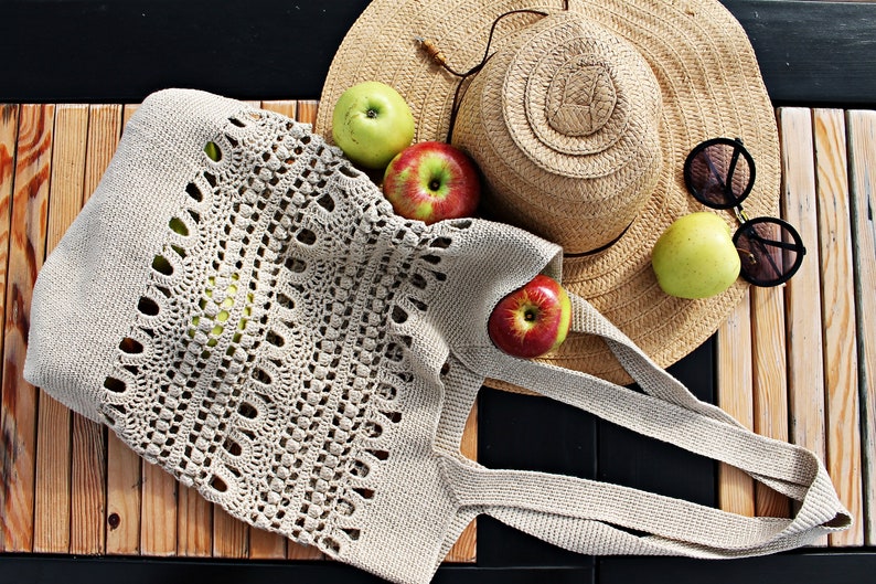 May include: A beige crocheted tote bag with a wide, open top and two long handles. The bag is lying on a wooden surface with a straw hat, sunglasses, and apples.