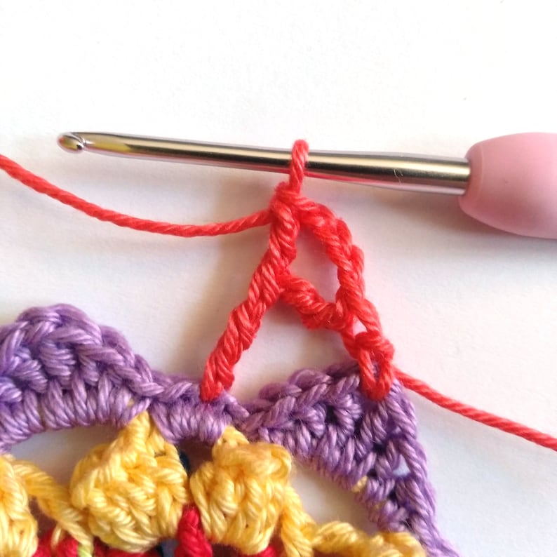 May include: Close-up of a crochet project with a pink crochet hook. The project is a multi-colored round doily with a red crochet stitch being worked on the edge.