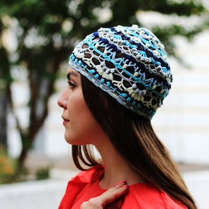 May include: A woman wearing a blue, white, and gray crocheted hat. The hat has a geometric pattern and is worn on her head.