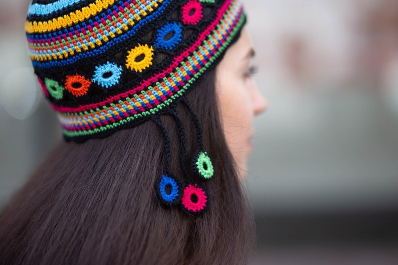 May include: A woman wearing a colorful crocheted hat with tassels. The hat has a black base with stripes of yellow, red, green, blue, and white. The hat is decorated with crocheted flowers in various colors.