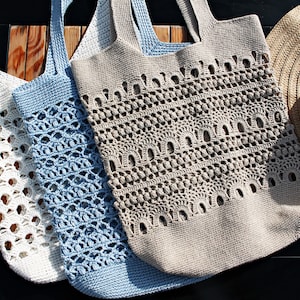 Könnte beinhalten: Drei gehäkelte Einkaufstaschen in Weiß, Blau und Beige. Die Taschen haben ein zartes Lochmuster und sind perfekt zum Tragen von Lebensmitteln oder anderen Gegenständen.