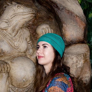 May include: A woman with long brown hair wearing a teal knitted hat stands in front of a large stone sculpture of a mythical creature. The sculpture is detailed with intricate carvings and appears to be a female figure.