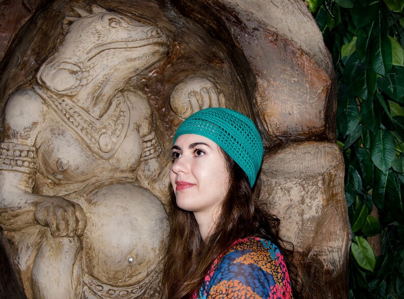 May include: A woman with long brown hair wearing a teal knitted hat stands in front of a large stone sculpture of a mythical creature. The sculpture is detailed with intricate carvings and appears to be a female figure.