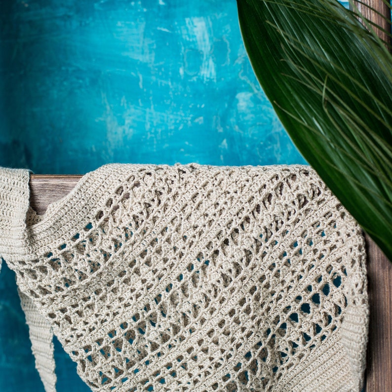 May include: A close-up of a beige crocheted tote bag with a diamond pattern. The bag is hanging on a wooden shelf against a blue background.