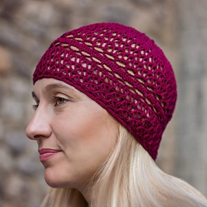 May include: A close-up of a woman wearing a burgundy crocheted beanie hat. The hat has a delicate openwork design.