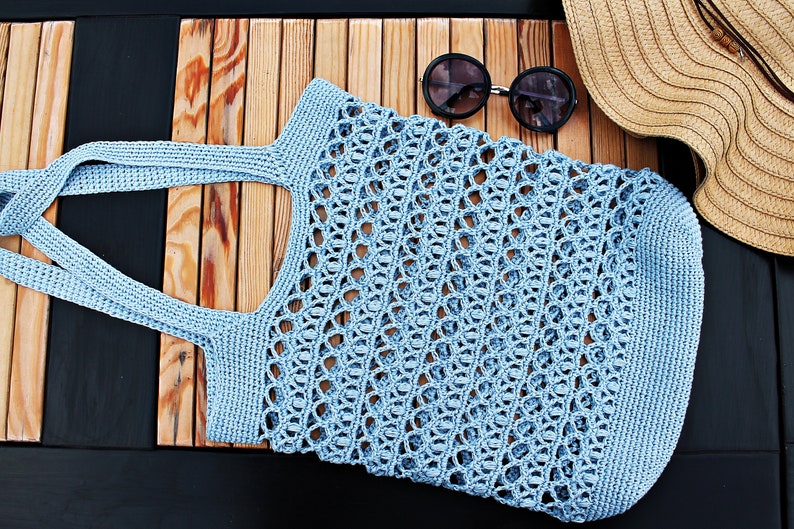 May include: A light blue crocheted tote bag with a woven pattern. The bag is lying on a wooden surface.