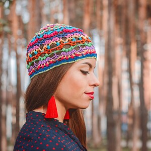 May include: A woman wearing a colorful crocheted hat with red tassel earrings. The hat has a rainbow pattern and is made with a variety of different colored yarns.