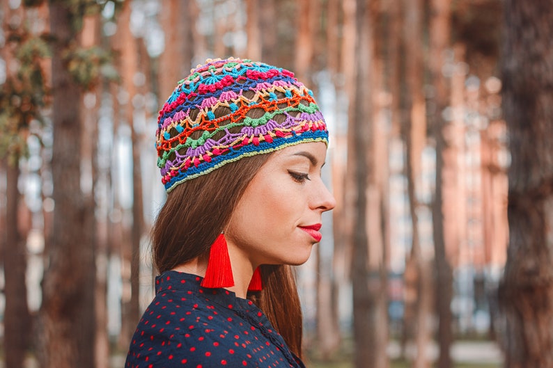May include: A woman wearing a colorful crocheted hat with red tassel earrings. The hat has a rainbow pattern and is made with a variety of different colored yarns.