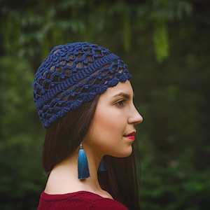 May include: A woman wearing a blue crocheted hat with long blue tassel earrings. The hat has a delicate openwork design. She is standing in front of a green background.