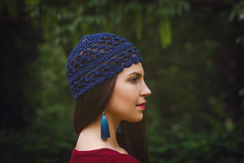 May include: A woman wearing a blue crocheted hat with long blue tassel earrings. The hat has a delicate openwork design. She is standing in front of a green background.