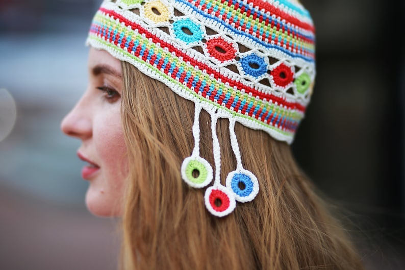 May include: A woman wearing a colorful crocheted hat with white tassels and colorful circles. The hat is made with a variety of colors, including red, blue, green, and yellow.
