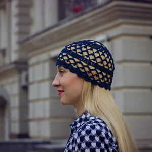 May include: A woman wearing a blue crocheted hat with a geometric pattern. The hat is worn on her head and covers her ears. She has long blonde hair and is wearing a black and white patterned shirt.
